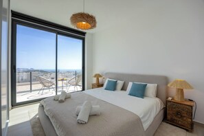 Een moderne slaapkamer met een groot bed, twee bedlampen, uitzicht op de stad door middel van kamerhoge ramen, en een balkon met stoelen.