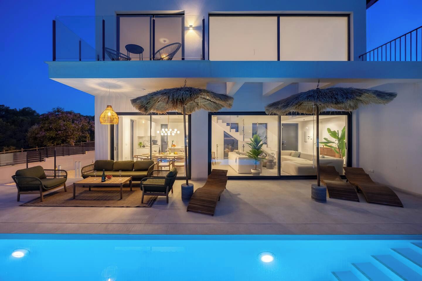 A modern two-story house with a swimming pool, outdoor seating, and a well-lit interior.