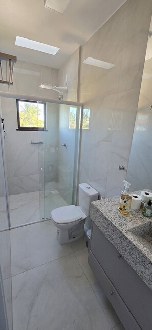 A modern bathroom with a glass shower enclosure, a toilet, and a granite countertop with toiletries. - Casa Duplex no Condominio Bella Arte (Aquiraz)