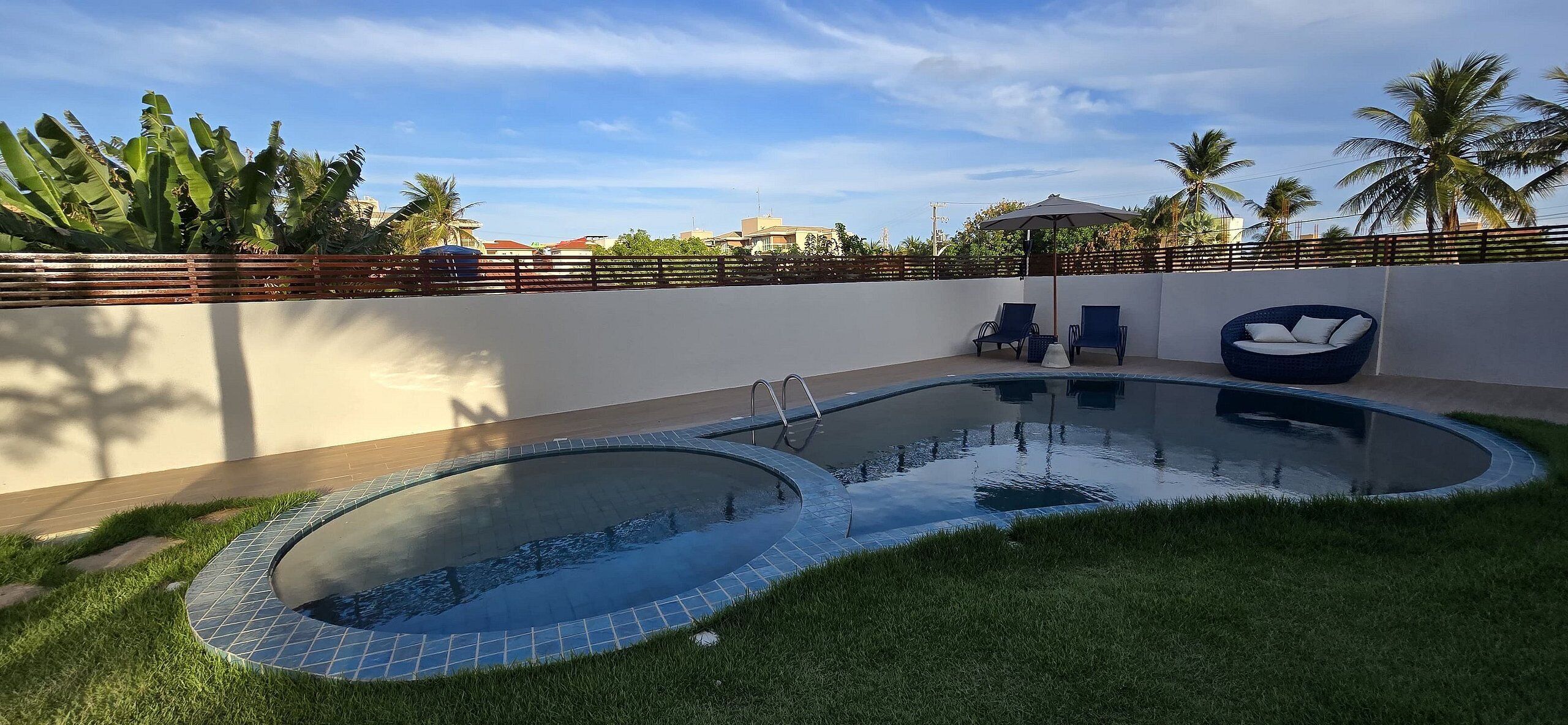 A swimming pool with a tiled edge, surrounded by grass, with two lounge chairs and a blue chair under a canopy.