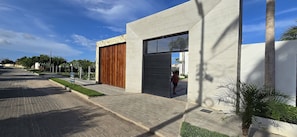 A modern building with a large wooden door and a metal shutter, a paved sidewalk, and a clear blue sky. - Casa Duplex no Condominio Bella Arte (Aquiraz)
