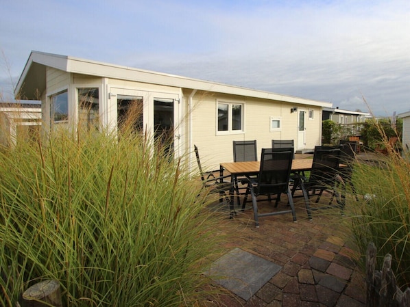 Eingeschossiges Haus mit einer Terrassenfläche, einem Esstisch mit Stühlen und einem kleinen Garten mit hohem Gras.