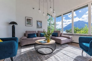 A modern living room with a sofa, two armchairs, a coffee table, and a view of mountains through large windows.