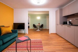 Een woonkamer met een bank, televisie en een klein tafeltje met een boek. Achtergrond toont een eethoek met stoelen en een tafel.