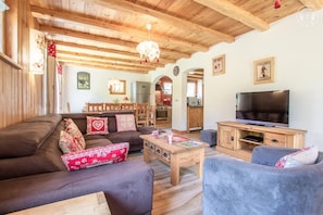 Ein gemütliches Wohnzimmer mit einer Couch, Sesseln, einem Couchtisch und einem Fernseher. Im Hintergrund ist eine Küche zu sehen.
