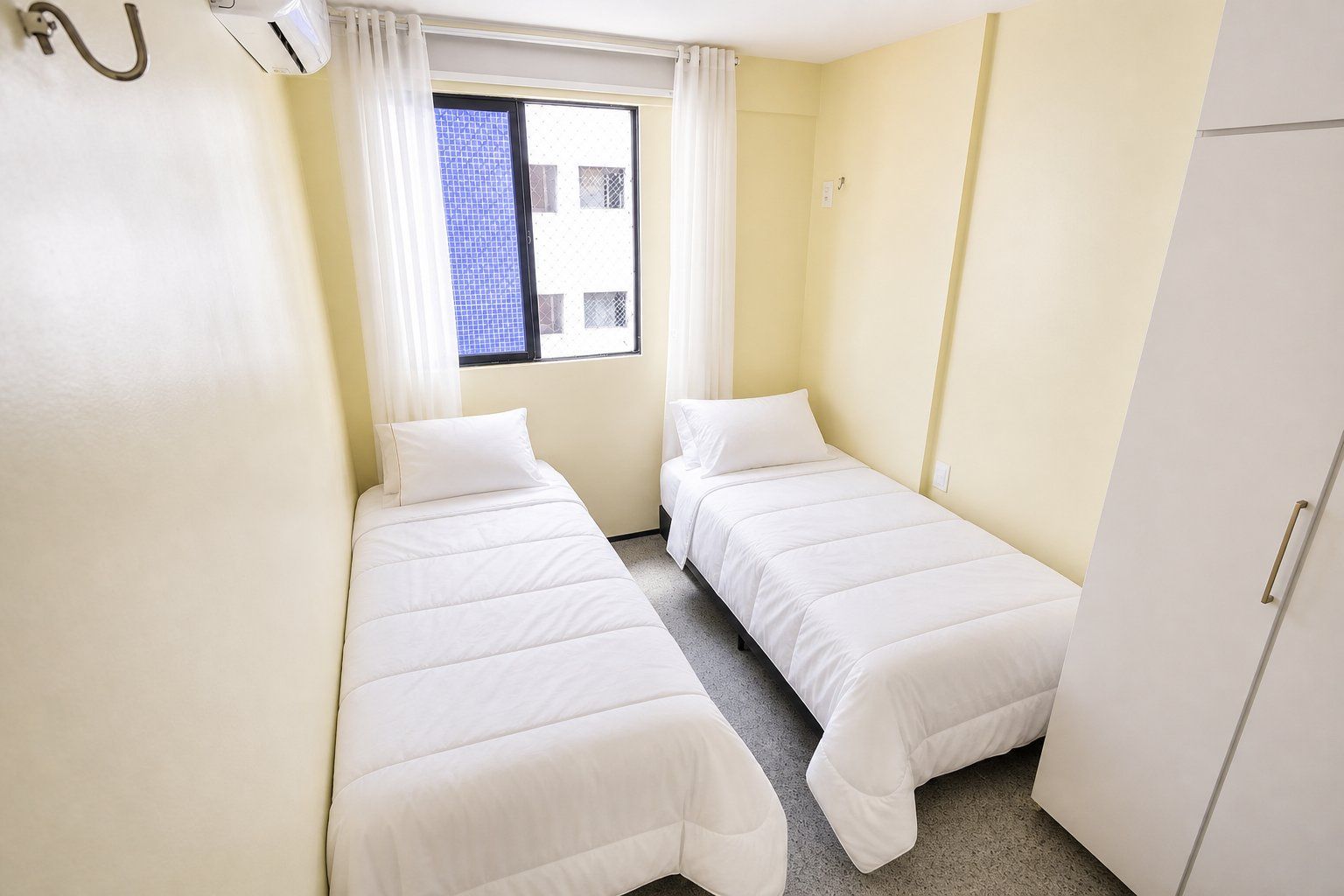 Quarto pequeno e limpo com duas camas de solteiro, janela com cortinas e um guarda-roupa branco.