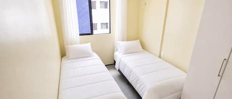 Quarto pequeno e limpo com duas camas de solteiro, janela com cortinas e um guarda-roupa branco.