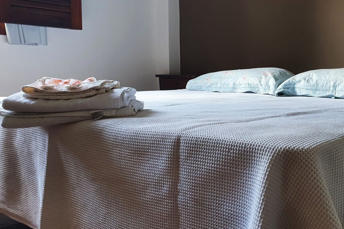 A neatly made bed with a white textured bedspread and two pillows, a stack of folded towels, and a dark nightstand.