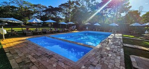 Una piscina rectangular con borde de azulejos, rodeada de sillas de descanso y sombrillas, con un sendero de ladrillo que la conduce.