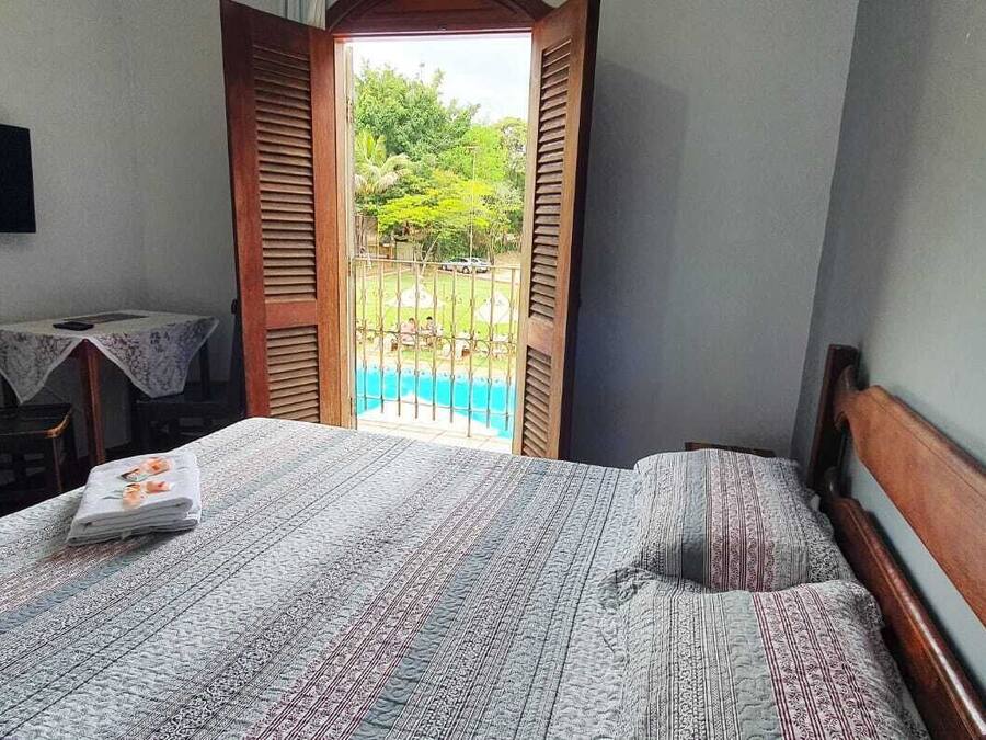 A bedroom with a bed, a TV mounted on the wall, a small table with a chair, and an open door leading to a balcony with a pool.