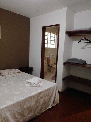 A bedroom with a bed, a bedside table, a hanging clothes rack, and a bathroom visible through a doorway. - Pousada Santana (Sabará)