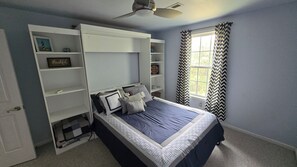 A bedroom with a bed, built-in shelving, a ceiling fan, a window with chevron curtains, and a 'NO WAKE ZONE' sign.
