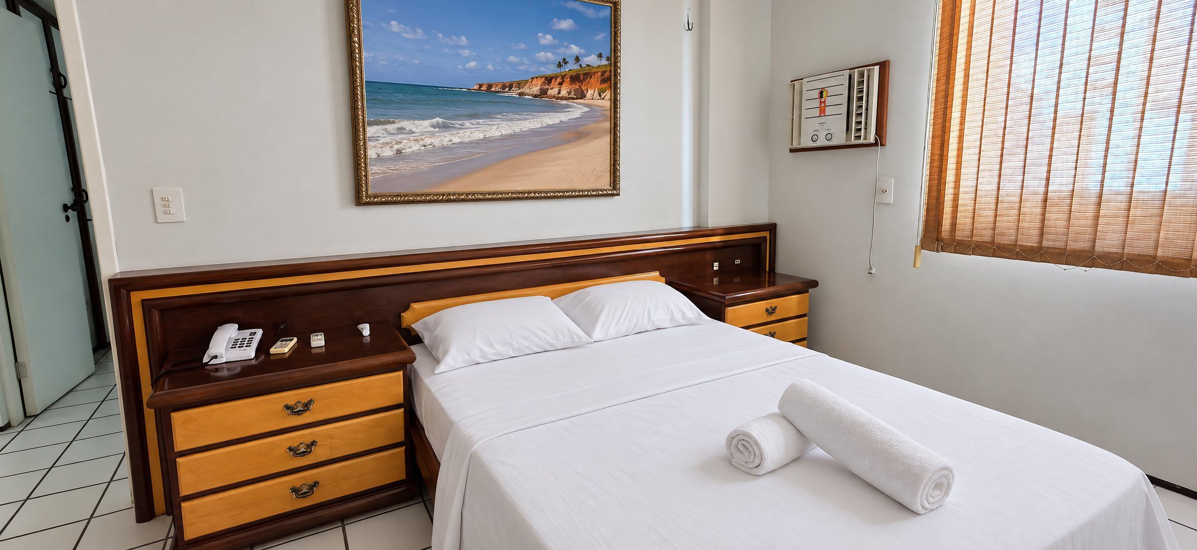 Ein Hotelzimmer mit einem Bett, einem Nachttisch mit Telefon, einem Beistelltisch, einem Strandbild und einem Thermostat an der Wand.