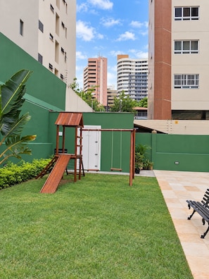 A grassy outdoor area with a swing set and a bench, surrounded by buildings. - Ape Familia 200 Metros da Feirinha Beira mar 502 (Fortaleza)