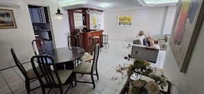 Een eetgedeelte met een ronde tafel en stoelen, een keuken op de achtergrond en een zithoek met een bank en een schilderij.