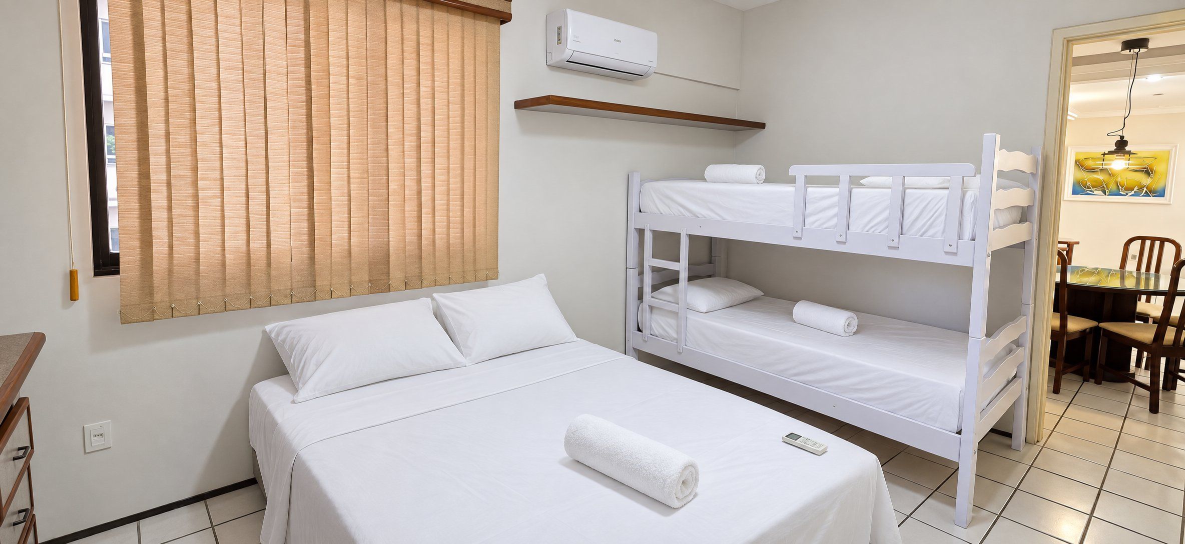 A hotel room with a bunk bed, a single bed, a window with blinds, an air conditioning unit, a small table with chairs, and a painting on the wall.
