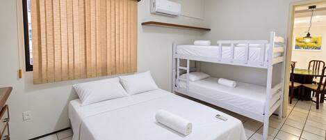 A hotel room with a bunk bed, a single bed, a window with blinds, an air conditioning unit, a small table with chairs, and a painting on the wall.