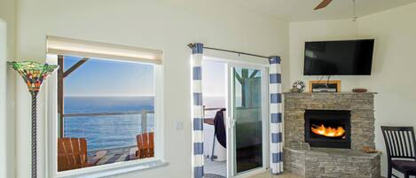 A living room with a stone fireplace, a television mounted above it, and a view of the ocean through a large window.