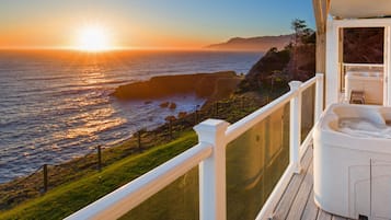 A balcony with a view of the ocean during sunset, a hot tub, and a wooden deck.