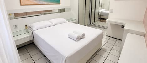 Quarto de hotel com cama, mesa de cabeceira, box de vidro para o chuveiro e um quadro com uma paisagem de praia.