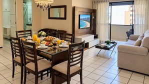 A modern living room with a dining area, a flat-screen TV mounted on the wall, and a large window with curtains. - Ape Familia 3 Quartos 200m da Feirinha Beira mar (Fortaleza)