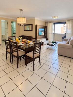 A modern living room with a dining area, a flat-screen TV mounted on the wall, and a large window with curtains. - Ape Familia 3 Quartos 200m da Feirinha Beira mar (Fortaleza)