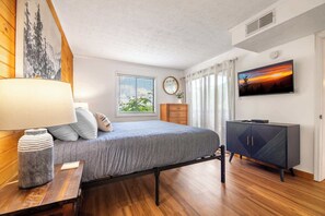 Ein Schlafzimmer mit Bett, Kommode, Fernseher, einem Fenster mit Jalousien und einem Bild an der Wand.