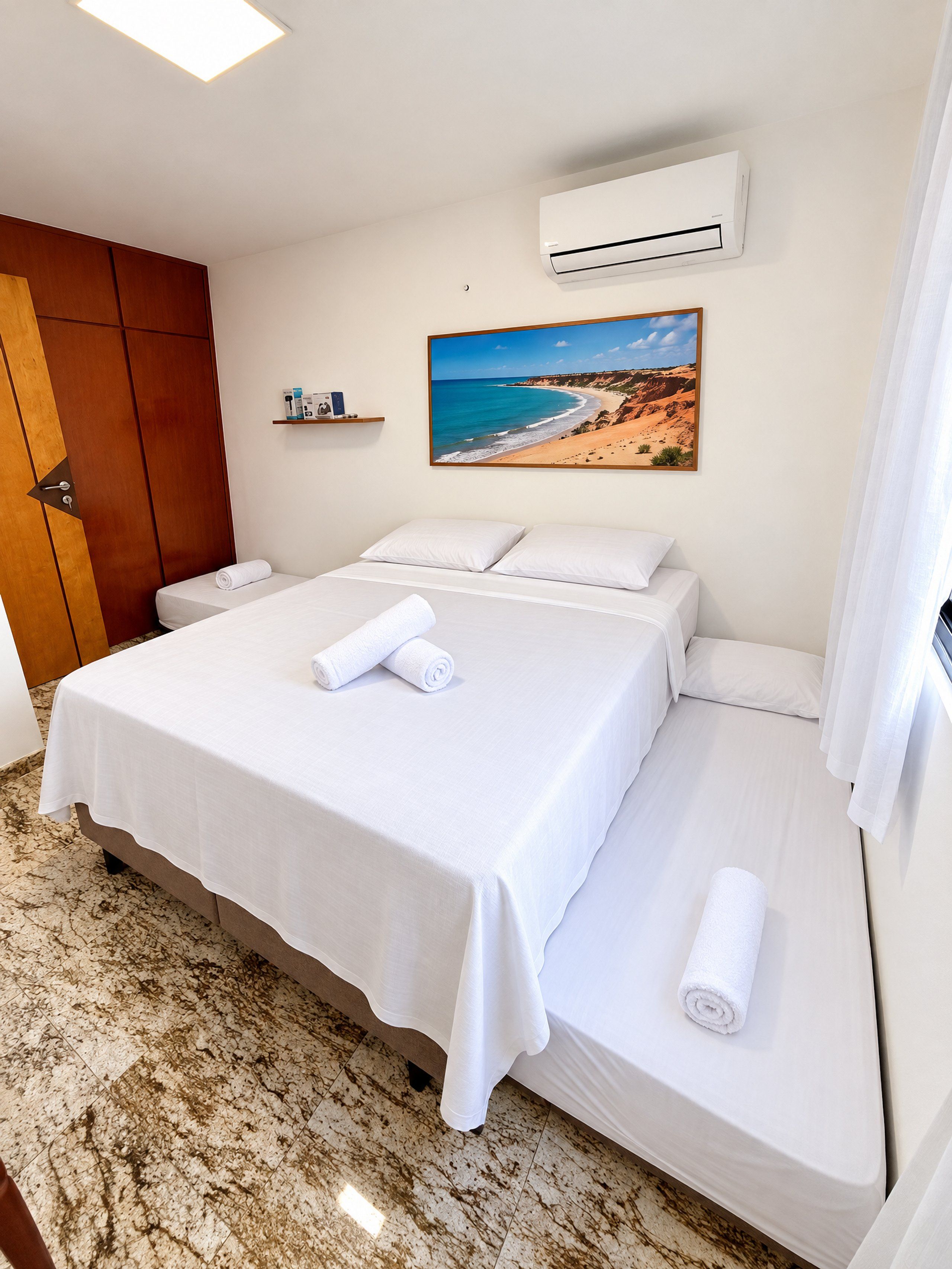 Quarto de hotel com cama, pintura de uma praia, unidade de ar condicionado e guarda-roupa de madeira.