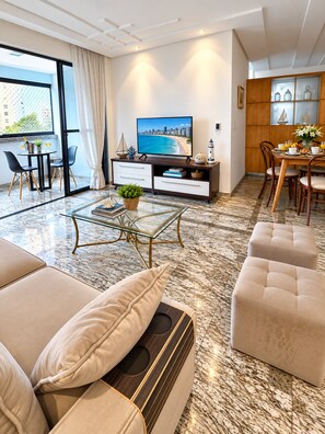 A modern living room with a flat-screen TV, a dining area with a wooden table and chairs, and a large window with a view of the city. - Apto Preferido Pelas Familias, 200m da Beira mar (Fortaleza)