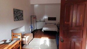 A bedroom with a bed, a bedside table, a chair, a small table with a cup and saucer, a fan, and a door.