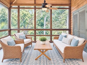 A room with a large window, wicker furniture, a ceiling fan, and a patterned rug.