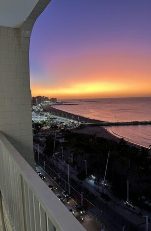 Ein Sonnenuntergangsblick von einem Balkon mit Blick auf einen Strand, eine Straße mit Fahrzeugen und eine ferne Stadtlandschaft.