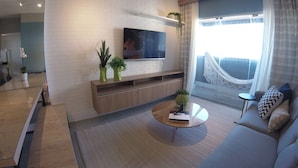 Un salon moderne avec un téléviseur à écran plat fixé sur un mur en briques blanches, un meuble de rangement en bois en dessous, une table basse ronde en verre, un canapé gris agrémenté de coussins à motifs et une grande fenêtre donnant sur un balcon.