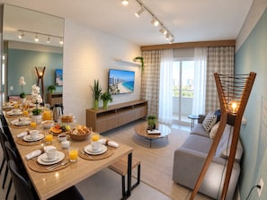 A modern living room with a dining area, a sofa, a TV mounted on a brick wall, and a balcony with curtains. - Beautiful Apartment With sea View 2 Bedrooms (Fortaleza)