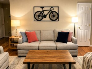 A living room with a sofa, a coffee table, a lamp, and a wall-mounted bicycle artwork. - Ozark Calling -- Ride-In Ride-Out MTB Basecamp, Premium Location (Bella Vista)