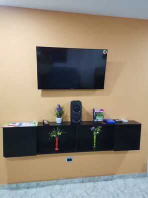 Un téléviseur fixé au mur, au-dessus d’un meuble noir servant de support pour l’équipement audio-vidéo, avec un haut-parleur, une télécommande et des objets décoratifs.