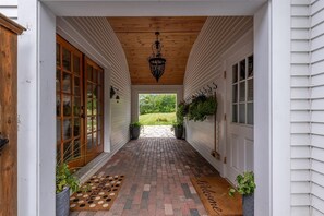A covered porch with a brick floor, a welcome mat, and potted plants.