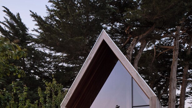 Una cabaña moderna de madera, con forma triangular y un frente de cristal, rodeada por un jardín y árboles altos.