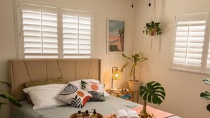 Un dormitorio con cama, ventilador de techo, mesita de noche con lámpara y dos ventanas con persianas.