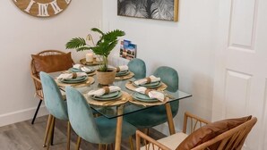 Un comedor con un reloj de madera redondo, un cuadro en blanco y negro de una hoja enmarco, y una mesa de vidrio con sillas color turquesa.