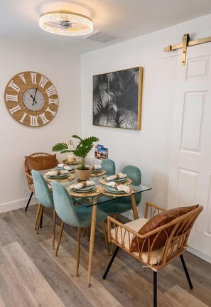 Un comedor con un reloj de madera redondo, un cuadro en blanco y negro de una hoja enmarco, y una mesa de vidrio con sillas color turquesa.