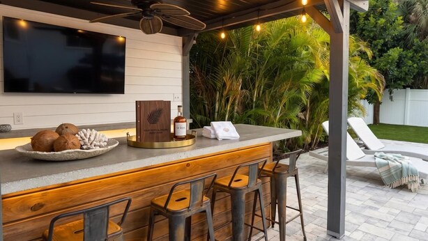Un área de bar al aire libre con mostrador de madera, taburetes, televisor y ventilador de techo.