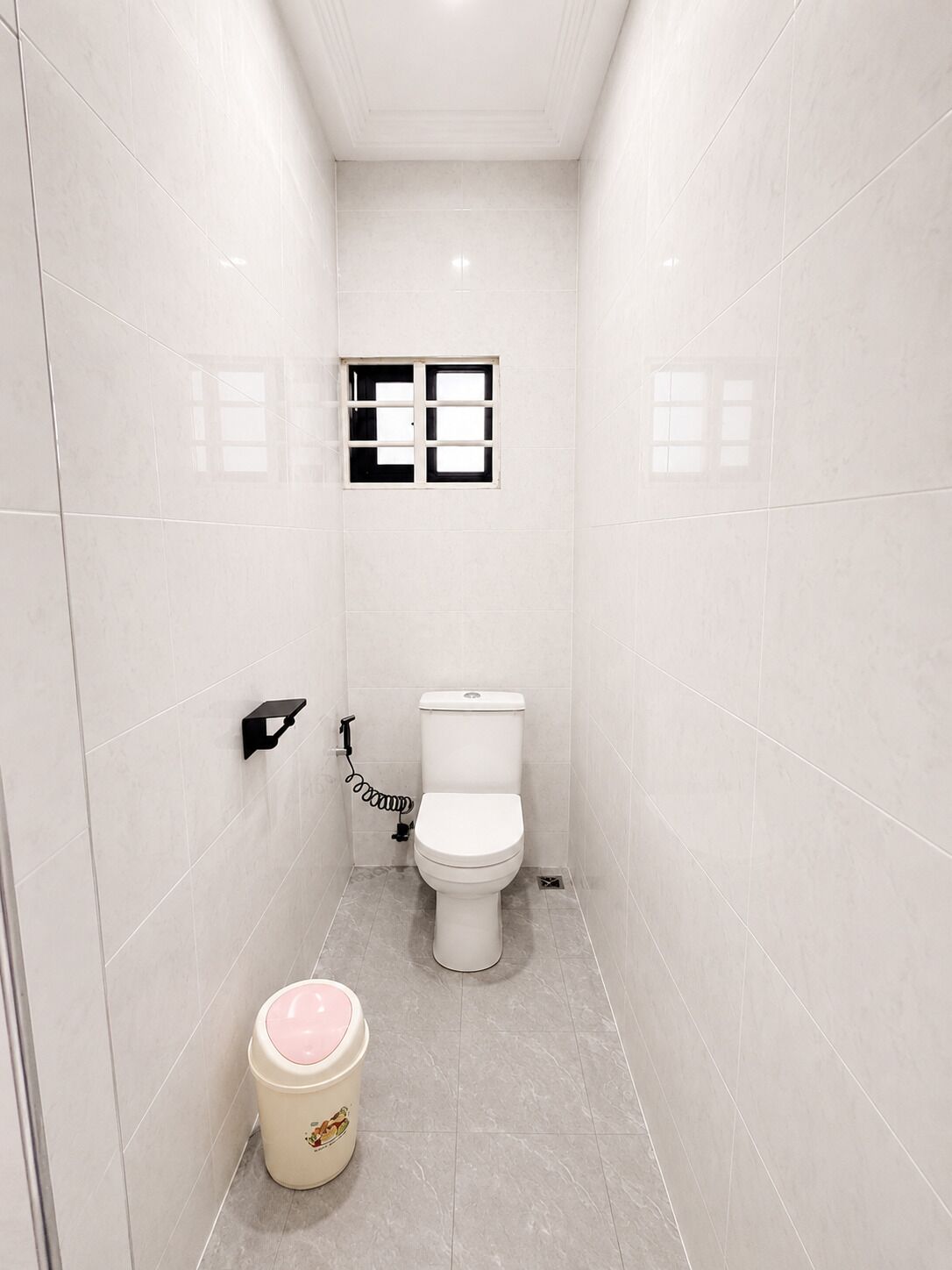 A narrow bathroom with white tiles, a toilet, a small window, and a trash bin.