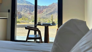 A hotel room with a bed, a wooden chair, a small table with a cup and saucer, and a view of mountains through large windows. - Casiraaaaa (Purmamarca)