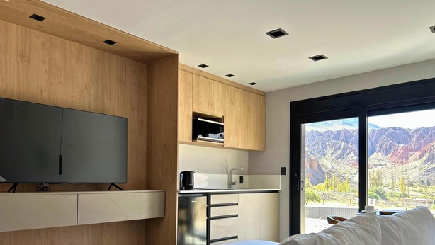A modern room with a flat-screen TV, wooden cabinetry, a kitchen area, and a large sliding glass door with a mountain view. - Casiraaaaa (Purmamarca)