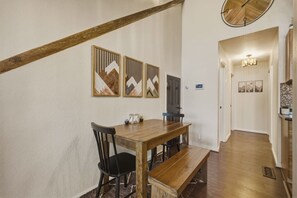 Uma área de refeições com mesa de madeira, cadeiras pretas e um banco. Há painéis decorativos na parede acima da mesa.