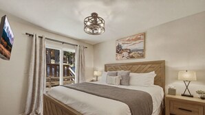 A bedroom with a bed, nightstands, a television, a painting, and a view of a balcony. - Cozy Alpine Retreat | BBQ, Views + Fireplace (Stateline)
