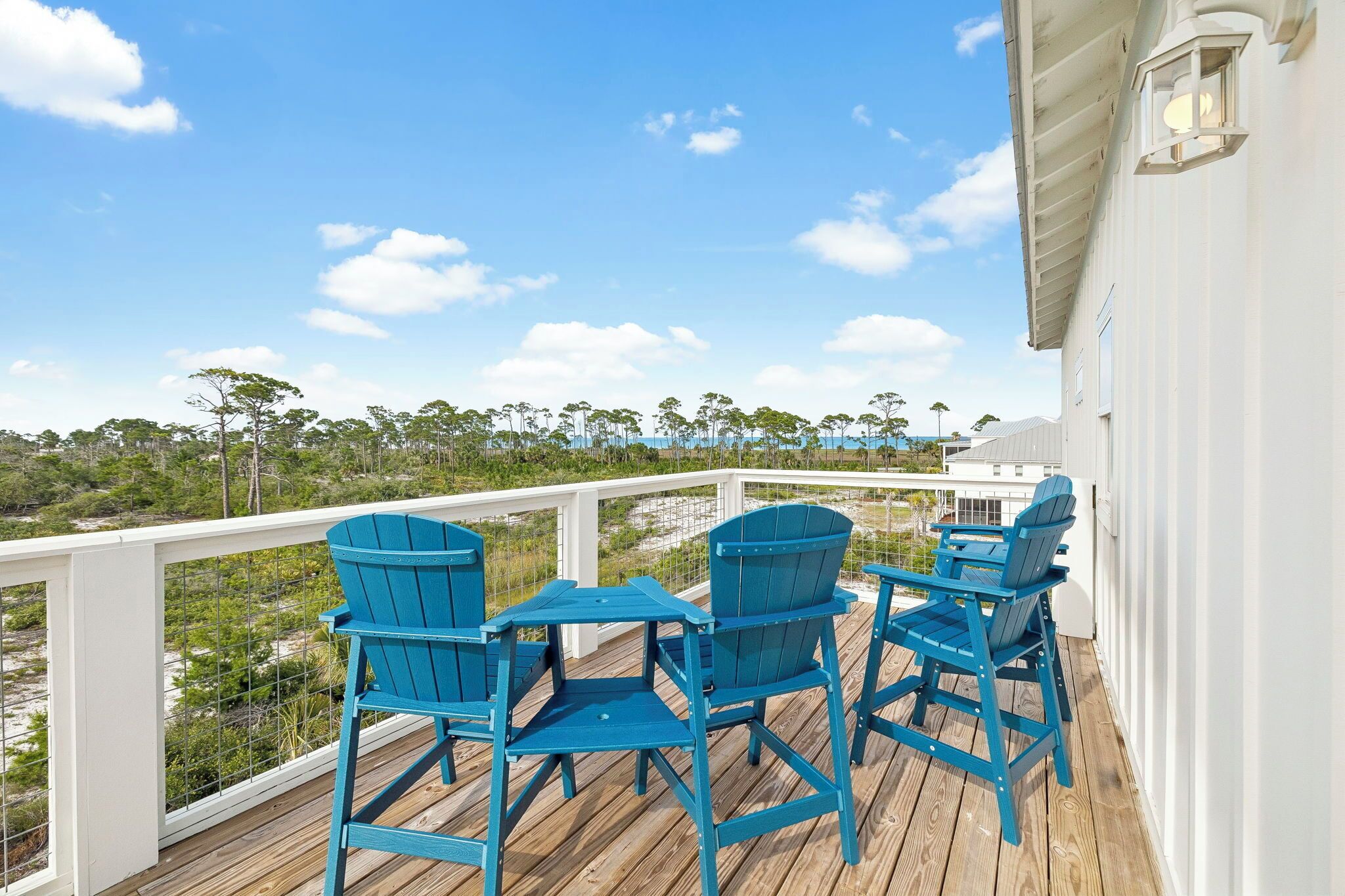 A wooden deck with blue Adirondack chairs and a white railing overlooking a beach and trees.