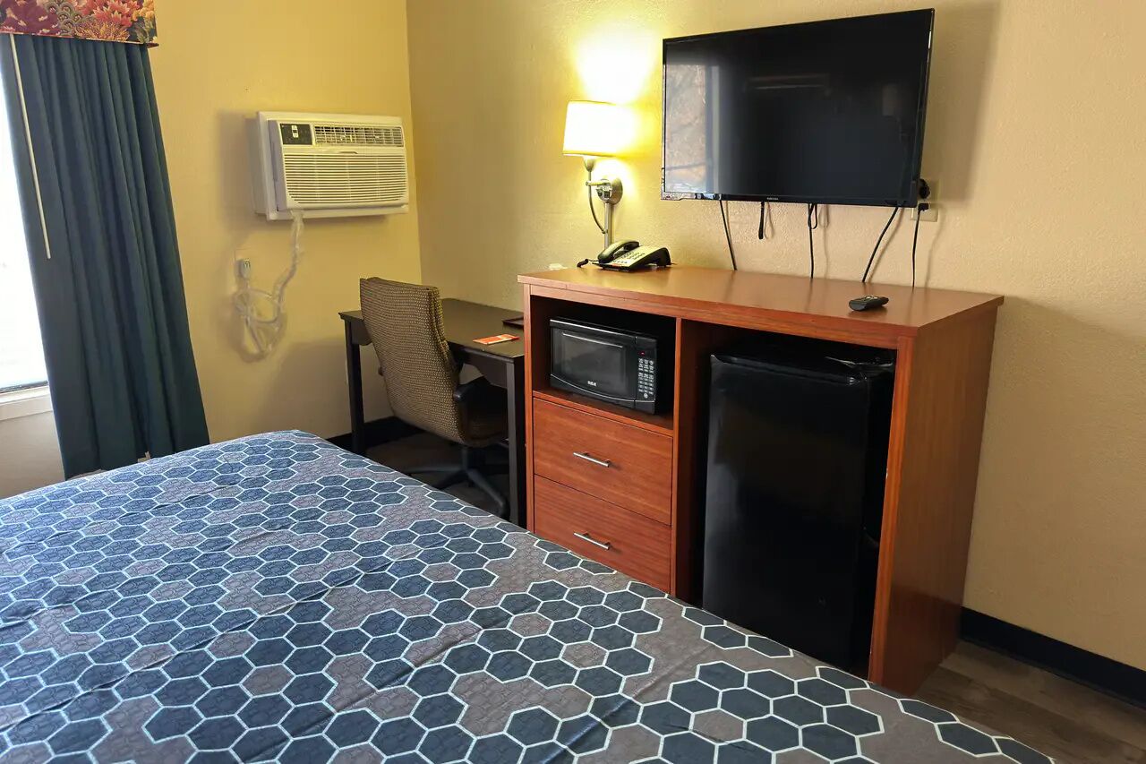 Een hotelkamer met een bed, een bureau met stoel, een magnetron, een koelkast, een televisie en een airconditioning.