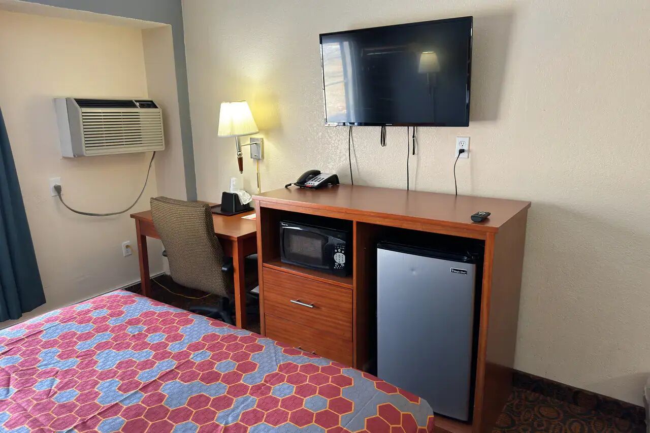 A hotel room with a bed, a desk with a telephone, a microwave, a refrigerator, an air conditioner, and a television.